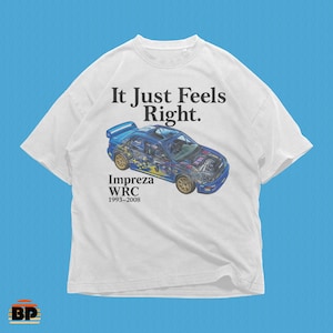 May include: White t-shirt featuring a blue Subaru Impreza WRC car graphic with cutaway details. The text "It Just Feels Right" is above the car, and "Impreza WRC 1993-2008" is below. The shirt has short sleeves.