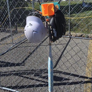 May include: A baseball helmet, glove, and bat are suspended on a fence. The helmet is white, the glove is black, and the bat is light blue with a white design. An orange and teal holder is attached to the fence.