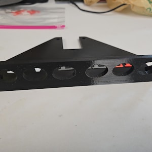 Puede incluir: Una estantería negra impresa en 3D con cinco aberturas circulares. La estantería tiene soportes triangulares y dos aberturas rectangulares en la parte superior. La estantería está diseñada para guardar objetos pequeños.