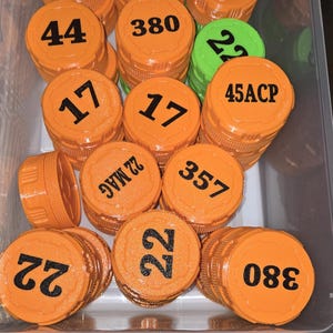May include: A selection of orange and green plastic caps, each marked with black numbers and text such as "17", "380", "9", "44", "22", "45ACP", and "357". The caps are stored in a clear plastic container.