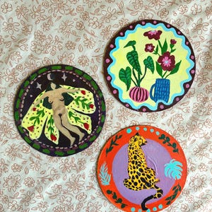 Puede incluir: Tres piezas decorativas redondas pintadas a mano. Una presenta una figura con alas amarillas, otra flores en un jarrón y la tercera un leopardo. Cada una tiene un esquema de color y diseño únicos, perfectas para la decoración de paredes.