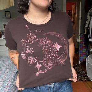 Könnte beinhalten: Ein braunes T-Shirt mit einer rosa Illustration eines Leoparden auf einem Mond, umgeben von Sternen und floralen Ranken. Das Oberteil hat einen Rundhalsausschnitt und kurze Ärmel. Das Model trägt blaue Jeansshorts.