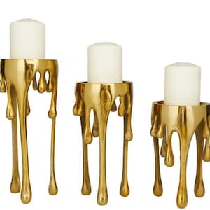 May include: Three gold-colored candle holders with white candles. The holders have a unique design, appearing as if the metal is melting or dripping. The candle holders are of varying heights, creating a tiered display.