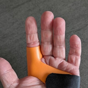 以下が含まれることがあります： 黒い布ストラップが付いたオレンジ色の指スプリント。スプリントは最初の3本の指を覆い、滑らかな素材で作られています。ストラップはスプリントを手に固定します。画像は医療またはサポートデバイスを示しています。