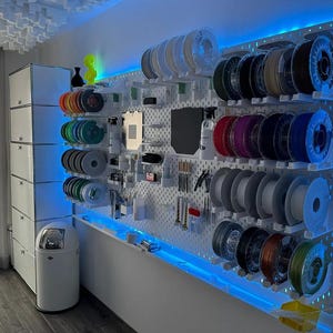 May include: A 3D printing filament storage system mounted on a wall. The system displays numerous filament spools in a variety of colours, arranged on a white pegboard. Blue LED lights illuminate the display, creating an organised workspace.