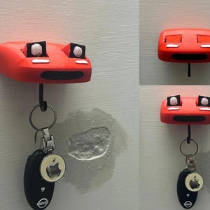 May include: A red key holder with a cartoon face design. The key holder has a black hook and is holding a car key with a silver Apple logo keychain. The key holder is mounted on a white wall.