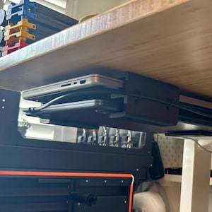 May include: A black under-desk laptop mount holding a silver laptop and a black tablet. The mount is attached to the underside of a light-colored wooden desk. Cables are neatly organized.
