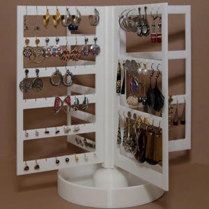 May include: A white, rotating earring display stand filled with various earrings. The stand has multiple panels with rows of holes to hang earrings. The earrings are in different styles, colors, and sizes, including hoops, dangles, and studs.