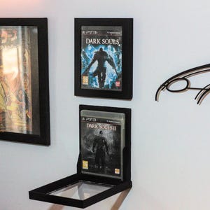 Op de afbeelding: Twee zwart omlijste game-hoesjes van Dark Souls en Dark Souls II, gemonteerd op een witte muur. De gamecovers hebben een dark fantasy-thema met een krijgersfiguur. De frames zijn rechthoekig en hebben een heldere voorkant.