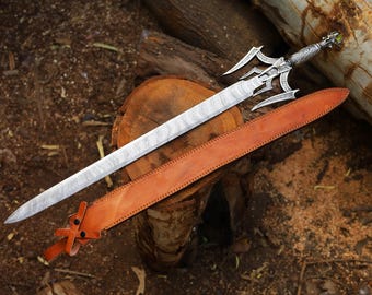 Puede incluir: Vista aérea de una espada con una hoja estampada y una empuñadura ornamentada, colocada sobre una superficie de madera. Una vaina de cuero marrón está colocada al lado. La empuñadura de la espada tiene una gema verde.