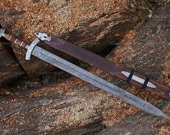 Puede incluir: Espada detallada con una hoja estampada y un mango de madera. Acompañada de una vaina de cuero marrón con grabados decorativos y detalles plateados. La espada descansa sobre una superficie de madera. La hoja mide unos 76 cm.