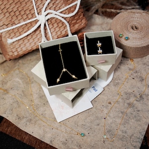 May include: An assortment of gold-colored jewelry, including necklaces and earrings, displayed in small, open boxes. One necklace features a cross pendant, while another has a small, round, blue gemstone. A gift box with a white ribbon is in the background.