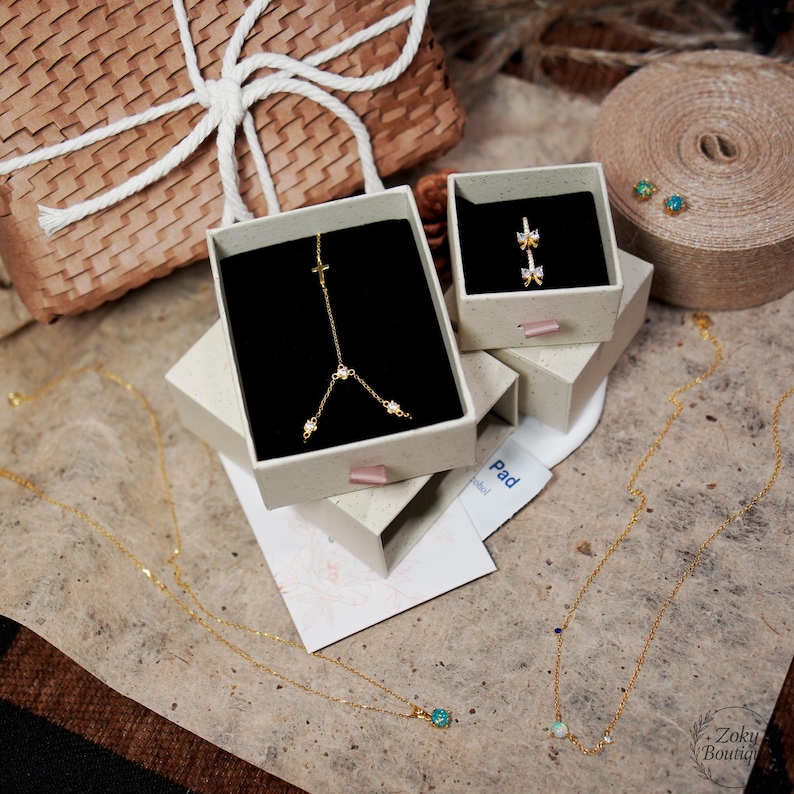May include: An assortment of gold-colored jewelry, including necklaces and earrings, displayed in small, open boxes. One necklace features a cross pendant, while another has a small, round, blue gemstone. A gift box with a white ribbon is in the background.
