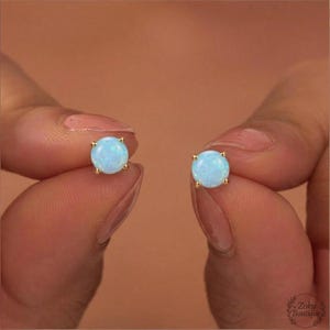 May include: Two gold stud earrings, each with a round, pale blue opal gemstone. The earrings are displayed against a warm, neutral backdrop, highlighting their subtle design and colour.