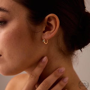 May include: Gold hoop earrings with a small, decorative floral design. The earrings are a warm gold color and feature a delicate, detailed flower with small, light-colored accents. The earrings are worn on the ear.