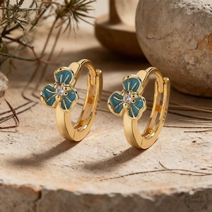May include: A pair of gold-colored hoop earrings. Each earring features a turquoise blue flower design with a small, clear gemstone in the center. The earrings are displayed on a textured surface.