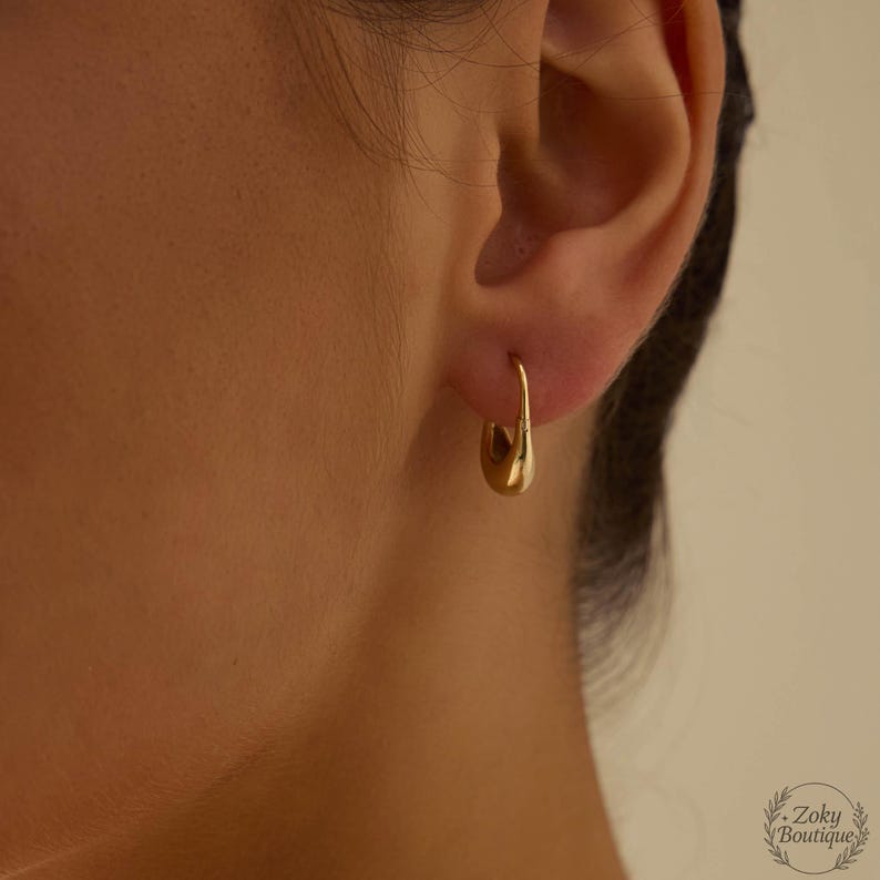 Puede incluir: Un solo pendiente de aro dorado se lleva en la oreja. El pendiente tiene un dise&ntilde;o liso y redondeado y un cierre seguro. El pendiente forma parte de una colecci&oacute;n de joyas.