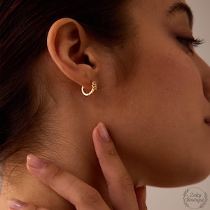 May include: A close-up of a gold hoop earring with a small, decorative charm. The earring is worn on the ear of a person. The earring is a simple, elegant design, perfect for everyday wear. The earring is made of a shiny, gold-colored metal.
