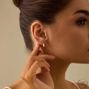 Puede incluir: Primer plano de una persona que lleva un par de pendientes de aro dorados peque&ntilde;os. Los pendientes tienen un dise&ntilde;o sencillo y pulido. La persona est&aacute; tocando uno de los pendientes con los dedos. El fondo es de un color suave y neutro.