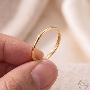 May include: A thin, polished gold-tone band ring with a smooth, rounded profile. The interior of the band features a clear 925 hallmark stamp, indicating sterling silver composition. The ring has a simple, minimalist design with a reflective surface.