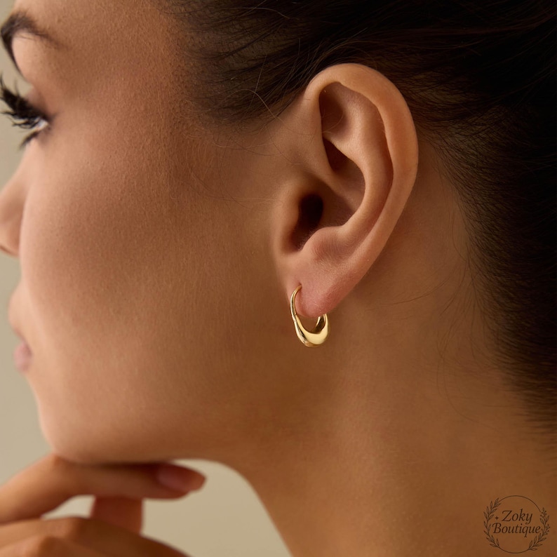Puede incluir: Primer plano de una oreja con un par de peque&ntilde;os pendientes de aro dorados. Los pendientes tienen un dise&ntilde;o liso y curvado. Los pendientes son un accesorio sencillo y elegante.