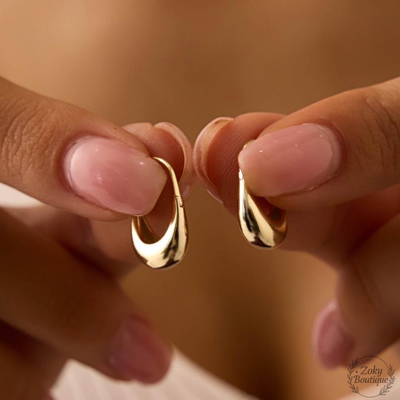 Puede incluir: Un par de pendientes dorados en forma de l&aacute;grima. Los pendientes son sostenidos por las manos de una persona, con el foco en la superficie pulida y reflectante de la joya. Los pendientes tienen un dise&ntilde;o liso y curvado.