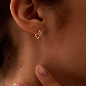 May include: Gold hoop earring with a small, three-leaf clover design. The clover has teal enamel and a small diamond accent. The earring is worn on the ear, with the ear and part of the face visible.