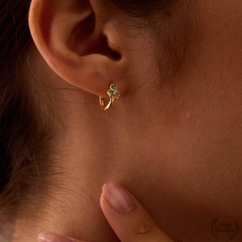 May include: Gold hoop earring with a small, three-leaf clover design. The clover has teal enamel and a small diamond accent. The earring is worn on the ear, with the ear and part of the face visible.