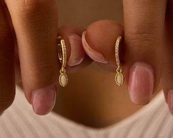 May include: Gold-colored hoop earrings with small white oval-shaped pendants. The hoops are encrusted with small, clear stones. The earrings are held up by a person's fingers. The earrings are from Zoky Boutique.