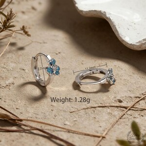 May include: Silver-tone hoop earrings with a blue flower design. The earrings are 11.2mm in diameter and 8.7mm in height. The weight of the earrings is 1.28g. The earrings are on a neutral-toned surface.