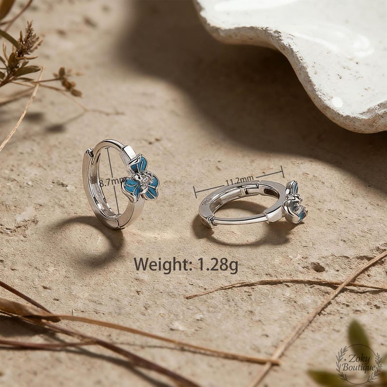 May include: Silver-tone hoop earrings with a blue flower design. The earrings are 11.2mm in diameter and 8.7mm in height. The weight of the earrings is 1.28g. The earrings are on a neutral-toned surface.