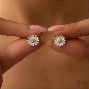 May include: A pair of daisy stud earrings. Each earring features a white flower with a gold-coloured centre. The earrings are held between fingers against a blurred background.