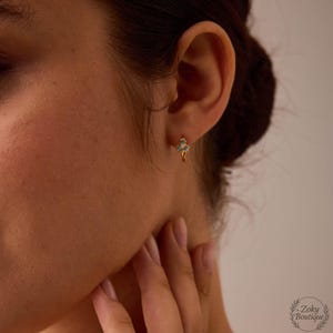 May include: Gold-colored hoop earrings with a small, three-leaf clover design. The clover is adorned with small, turquoise-colored stones. The earrings are worn on the ear of a person.