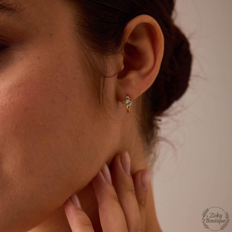 May include: Gold-colored hoop earrings with a small, three-leaf clover design. The clover is adorned with small, turquoise-colored stones. The earrings are worn on the ear of a person.
