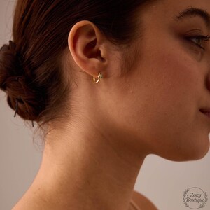 May include: A close-up of a person's ear wearing a small, gold hoop earring. The earring features a small, decorative element with a touch of color. The person's hair is pulled back, and the background is softly blurred.