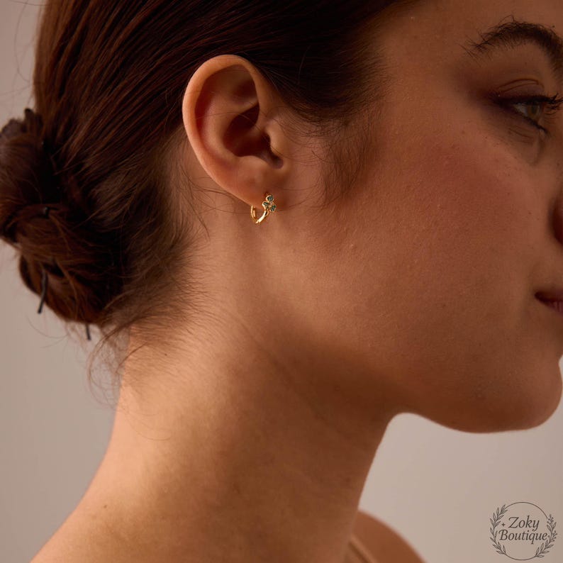 May include: A close-up of a person's ear wearing a small, gold hoop earring. The earring features a small, decorative element with a touch of color. The person's hair is pulled back, and the background is softly blurred.