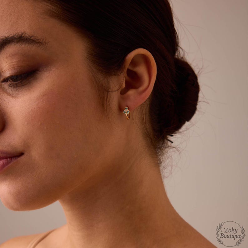 May include: A close-up of a person's ear wearing a small gold hoop earring with a floral design. The earring features small, sparkling stones. The background is a soft, neutral color, highlighting the jewelry.