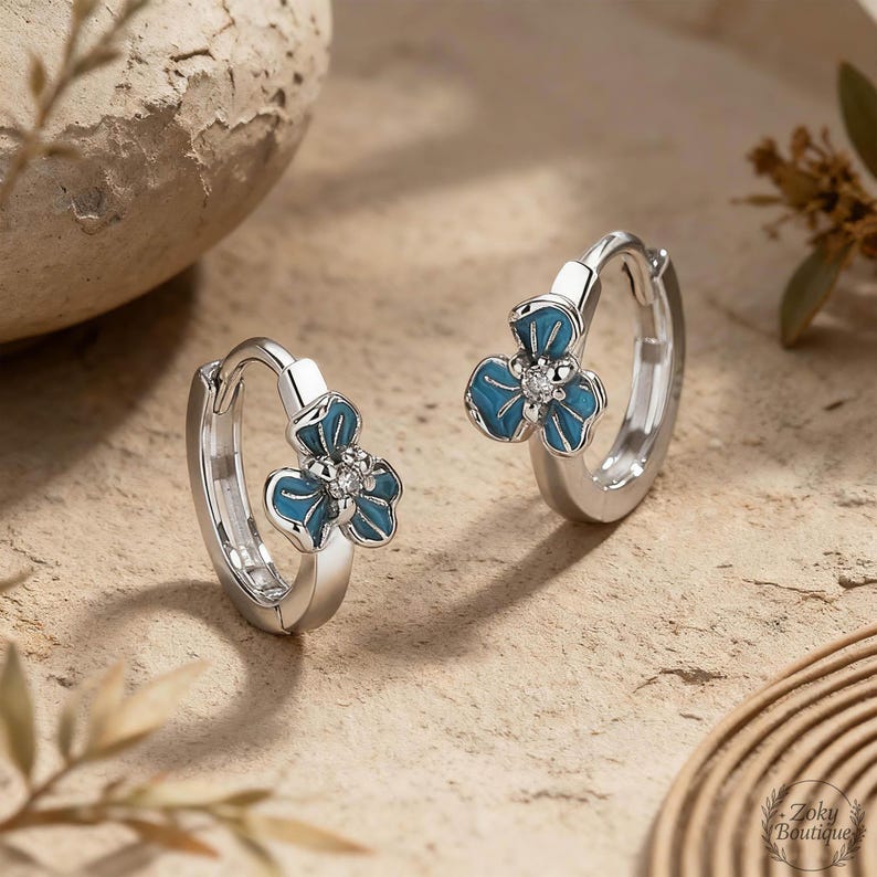 May include: A pair of silver hoop earrings, each adorned with a blue flower design. The flowers have three petals and a small, clear gem in the center. The earrings are displayed on a textured, light brown surface.
