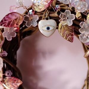 May include: A decorative wreath made of brown twisted twigs adorned with translucent pink and gold-toned leaf charms, small clear crystal beads, and pale pink flower accents. A central white heart-shaped charm features a blue evil eye design.