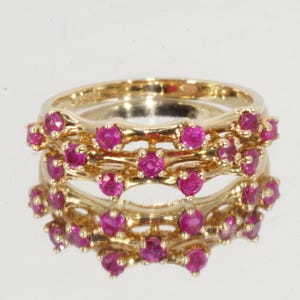 May include: A gold ring featuring a multi-band design with a series of small, round, vibrant pink rubies set along the metal structure. The ring has a polished finish and is captured with a clear reflection on the surface beneath it.