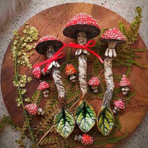 Puede incluir: Una colección de obras de arte artesanales con temática de setas. La exhibición incluye tres setas grandes con sombreros rojos y puntos blancos, atadas con cintas rojas, y varias setas más pequeñas. Hojas verdes y helechos están dispuestos sobre una bandeja de madera.