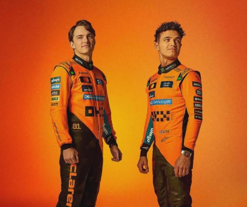 May include: Two people wearing orange and black racing suits with sponsor logos. The suits have long sleeves and are paired with black pants. The background is a gradient of orange.