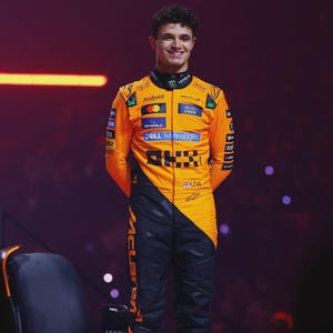 May include: A smiling person wearing a bright orange and black racing suit. The suit has sponsor logos and the word "McLaren" printed on the leg. The person is standing on a stage with a dark background.