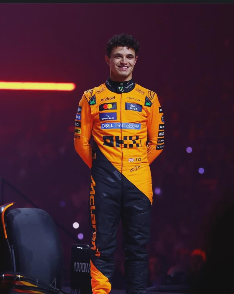 May include: A smiling person wearing a bright orange and black racing suit. The suit has sponsor logos and the word "McLaren" printed on the leg. The person is standing on a stage with a dark background.