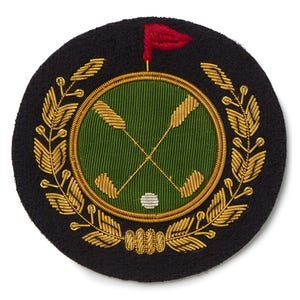 May include: A round, black embroidered patch with a golf theme. The design features crossed golf clubs, a golf ball, and a red flag on a green background, surrounded by a gold laurel wreath. The patch is likely for apparel or accessories.