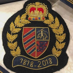 Puede incluir: Parche bordado negro con diseño de escudo. El escudo presenta un escudo rojo y azul, una corona dorada y los años 1878-2018. Hojas de laurel doradas y hilo dorado adornan el diseño.