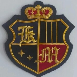 Puede incluir: Parche bordado en forma de escudo con una corona. El parche está dividido en secciones negras y rojas, con letras bordadas en oro y un borde dorado. La corona es dorada con detalles rojos.