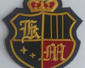 May include: Embroidered patch in a shield shape with a crown. The patch is divided into black and red sections, with gold embroidered letters and a gold border. The crown is gold with red accents.