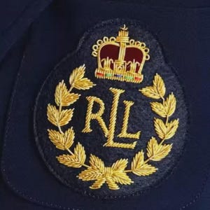 以下が含まれることがあります： 金色の王冠、月桂樹の葉、そして「RL」の文字が刺繍された、濃いネイビーブルーのパッチのクローズアップ。 楕円形のパッチは、おそらく衣服である濃いネイビーブルーの生地に縫い付けられています。