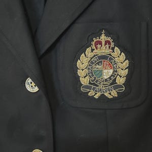 Pode incluir: Um blazer preto com um botão dourado e um emblema bordado proeminente no bolso do peito. O brasão exibe uma coroa, folhas de louro e o texto Ralph Lauren em fios dourado, vermelho, verde e branco sobre um fundo preto.